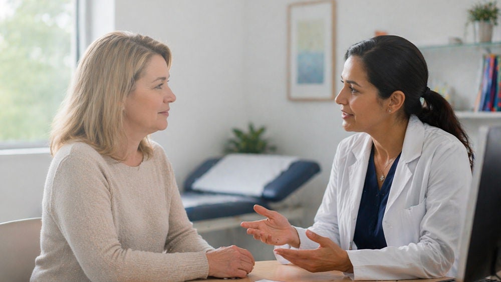 A female doctor in a white coat speaks with a middle-aged woman in a medical office. They sit across from each other at a desk, engaged in a focused conversation.