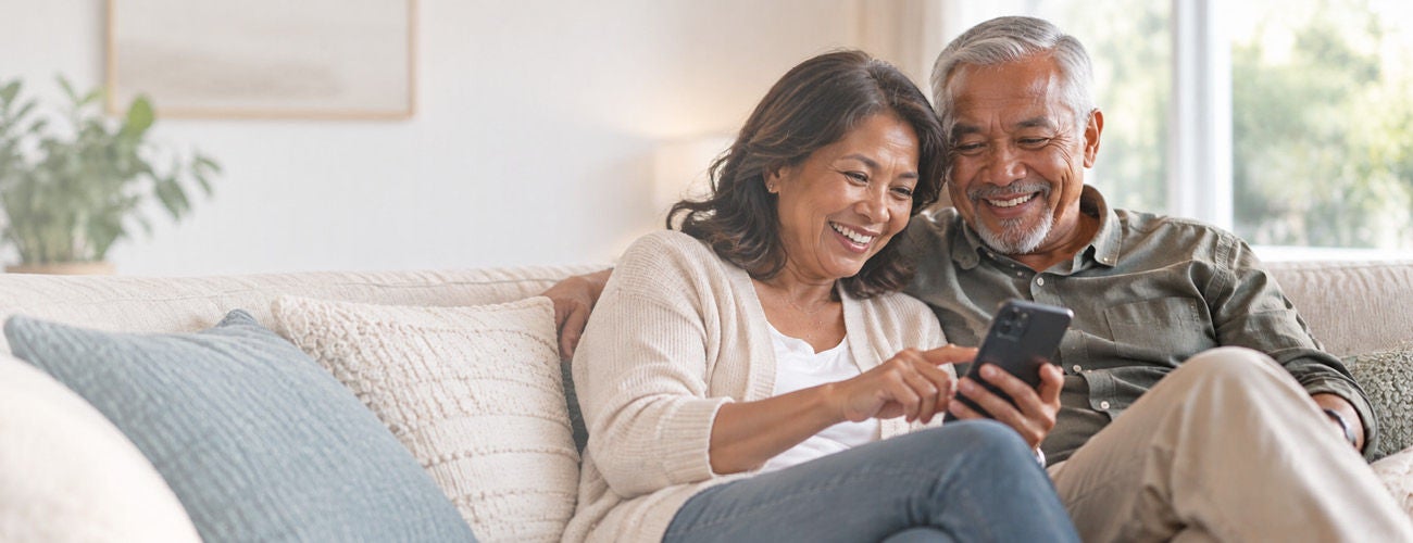 A smiling mature couple sits closely together on a comfortable sofa, looking at and interacting with a smartphone.