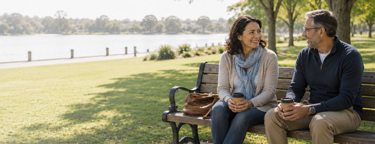 A middle-aged man and woman walk side by side along a paved path beside a calm waterfront. They appear relaxed and engaged in conversation, enjoying a sunny day outdoors. 
