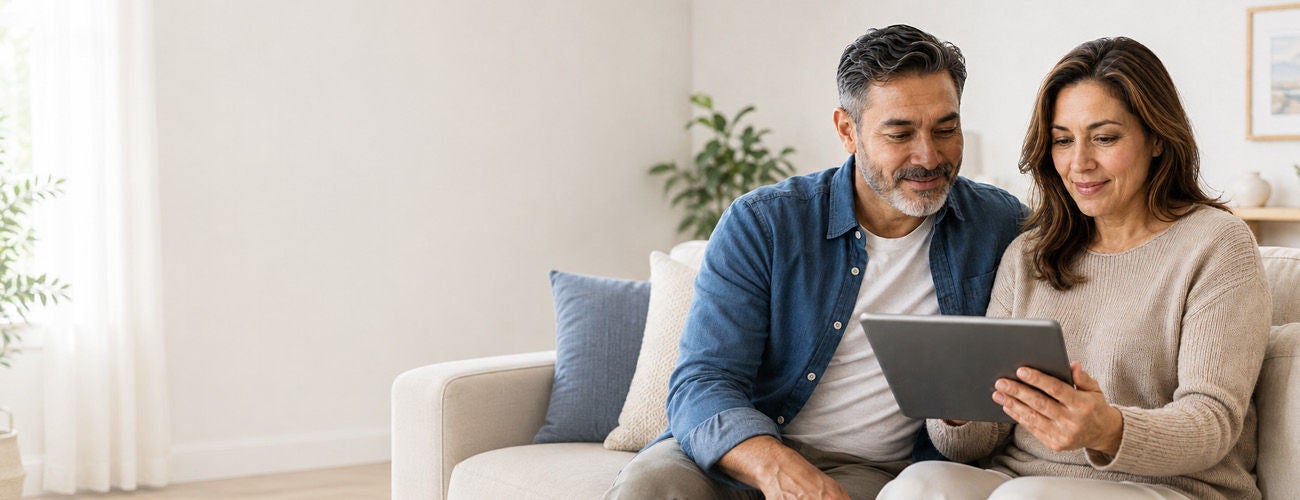 A middle-aged couple sits together on a light-colored sofa, looking at a digital tablet in the woman's hands.
