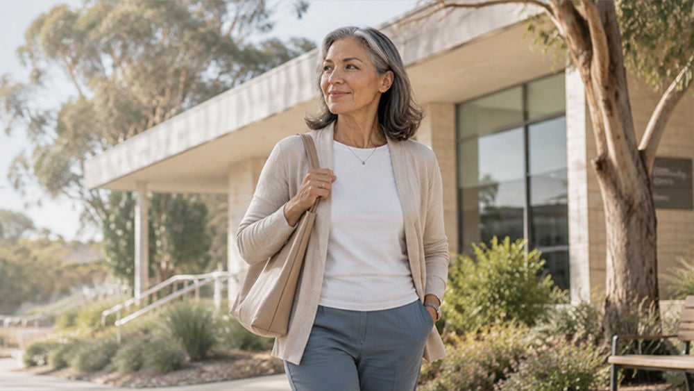 An older woman with gray hair walks confidently outside a modern building, carrying a beige shoulder bag.