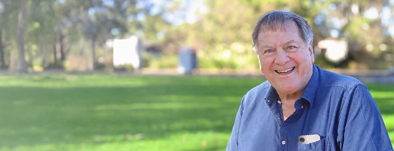 An older man sits outdoors, smiling warmly at the camera.
