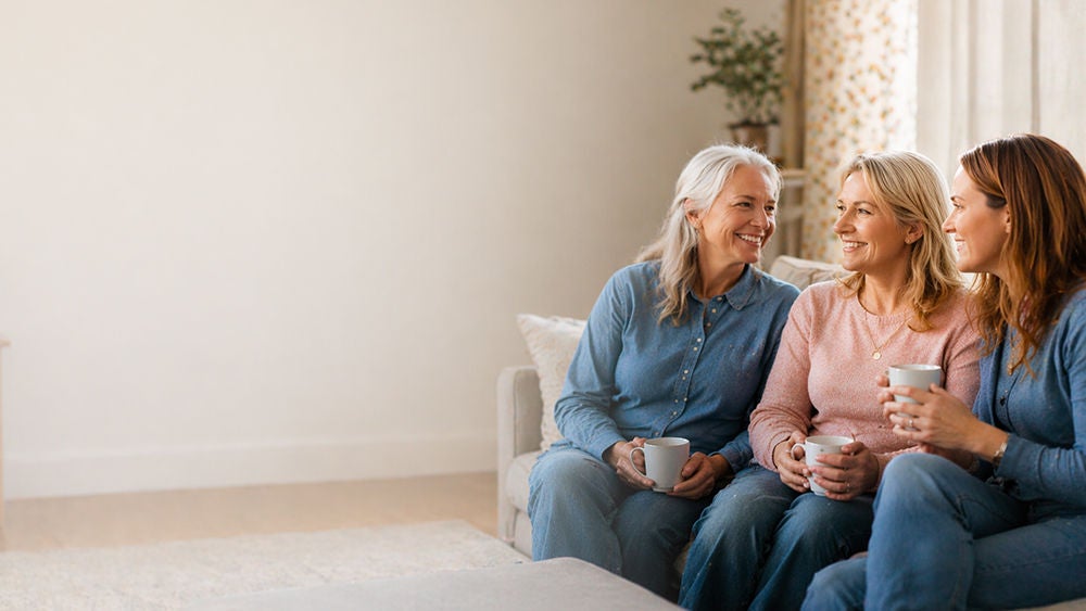 hree women sit closely together on a sofa, smiling and chatting while holding mugs.