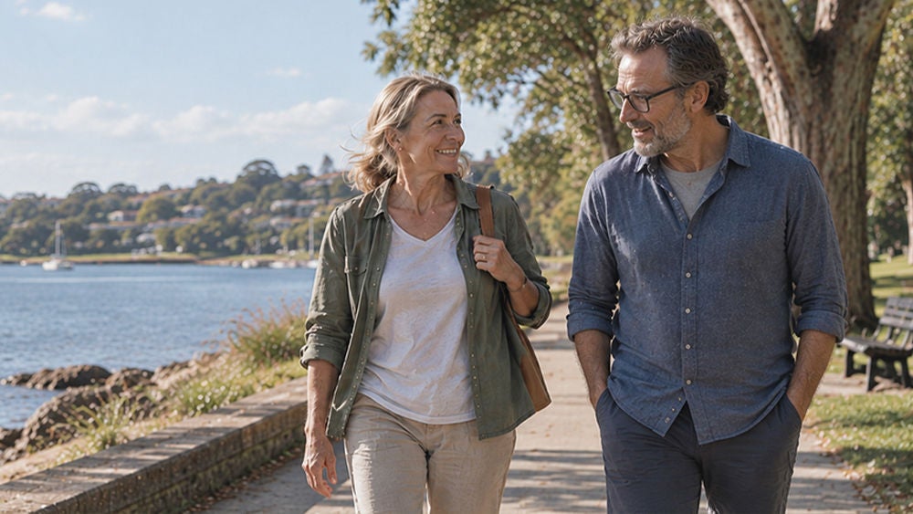A middle-aged woman and man walk side by side along a paved path beside a waterfront. 