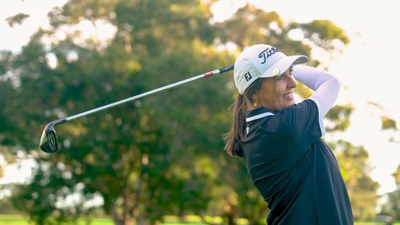 A woman golfer is captured mid-swing on a sunny day, smiling as she follows through with her club. 