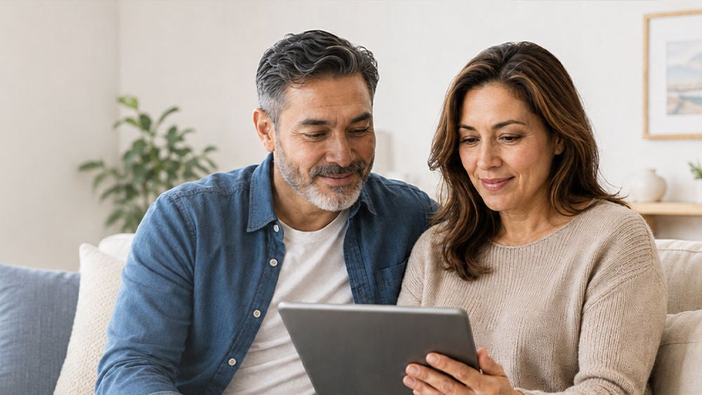 A middle-aged couple sits closely together on a comfortable sofa, looking at a digital tablet in their hands.
