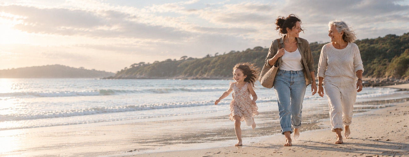 A young woman, an older woman, and a child walk together along a sandy beach near the shoreline.
